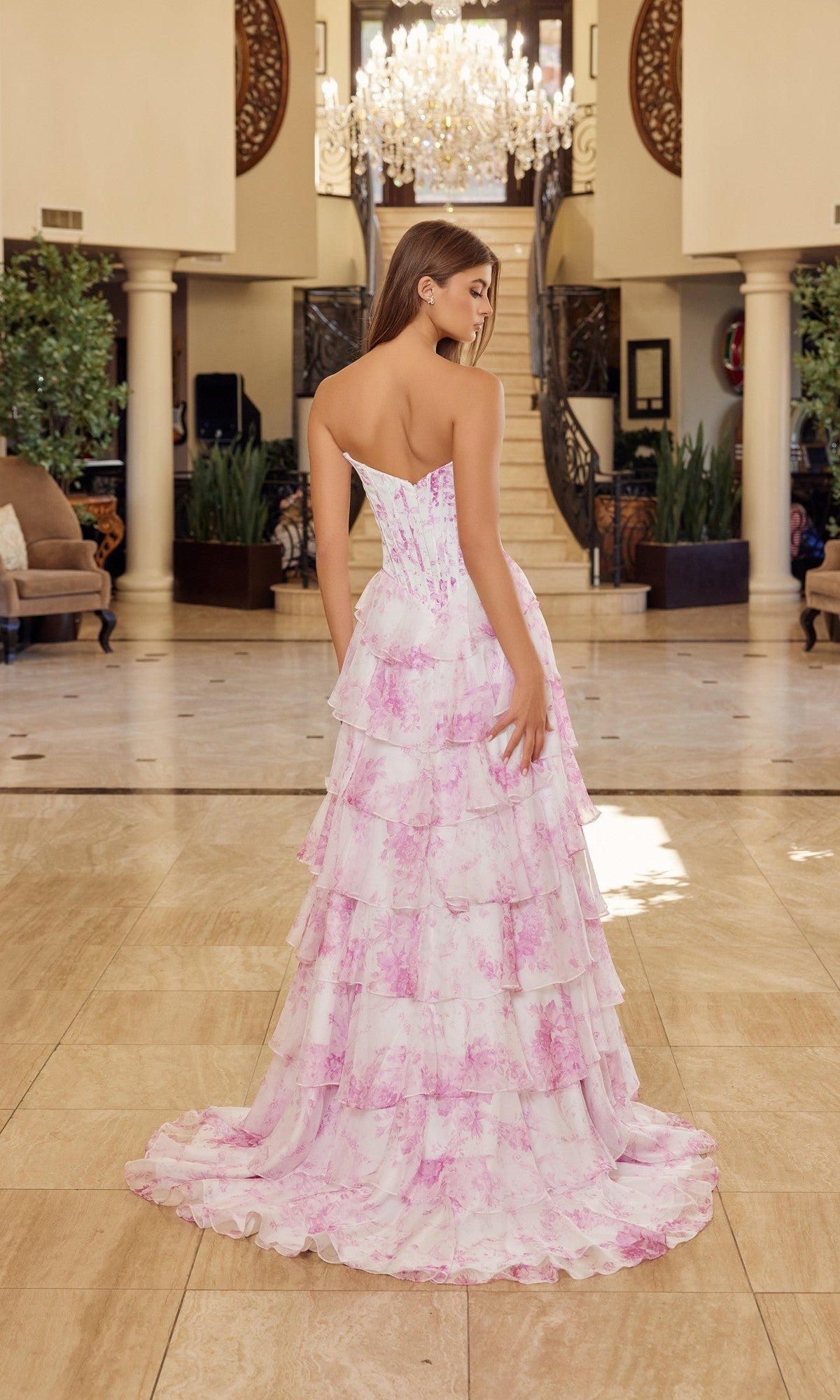 A woman stands indoors on marble flooring, facing away from the camera toward a staircase and chandelier, wearing the Nox Anabel A1752—a long prom dress with a strapless satin corset bodice and floor-length A-line skirt.